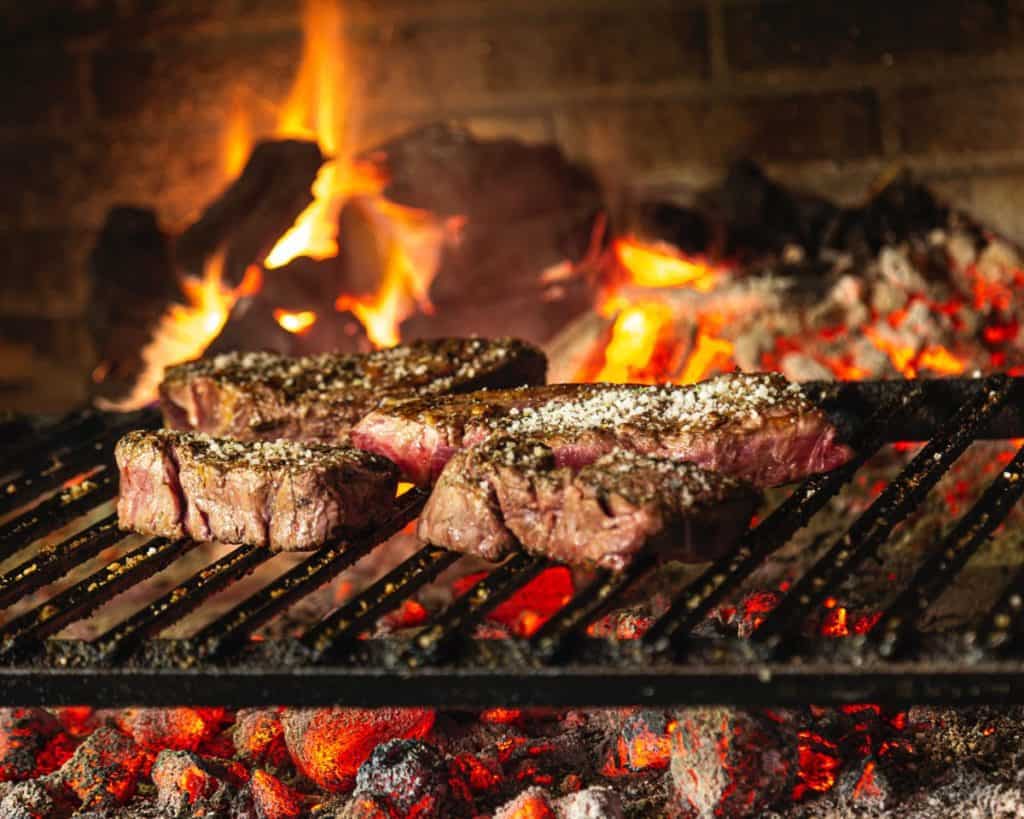 Seasoned steaks cooking on a metal rack directly over an open fireplace fire