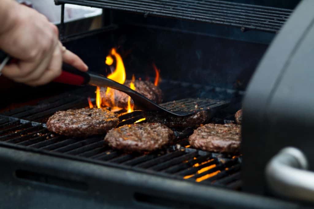 A hand using tongs to flip burgers over open flame on a gas grill
