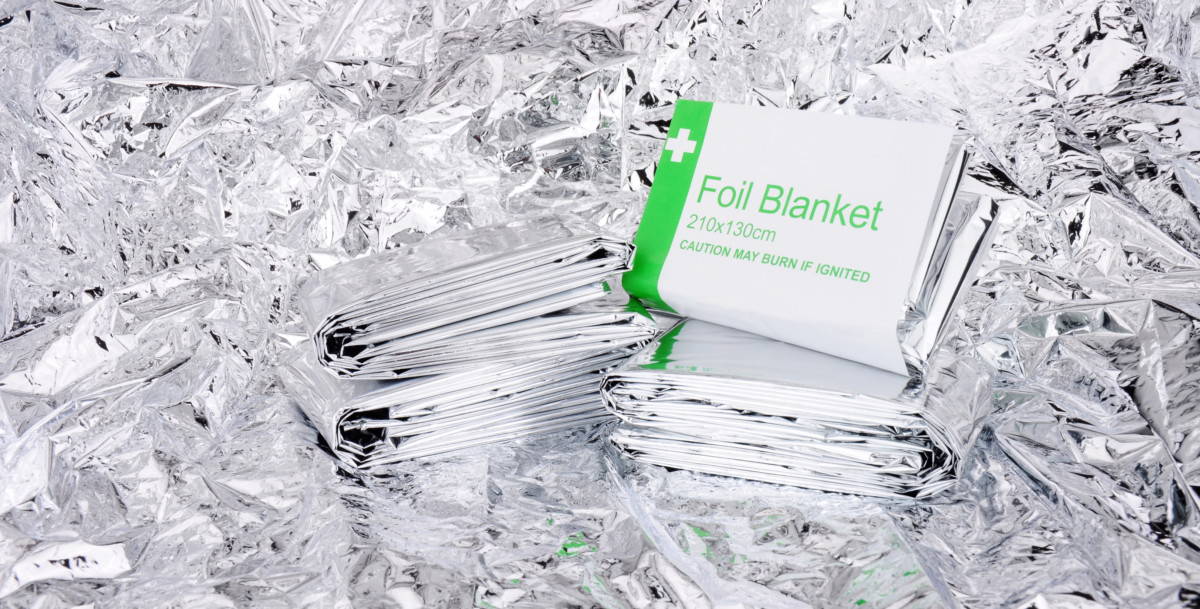 Three foil emergency blankets folded in their wrappers, resting on a crumpled sheet of silver mylar