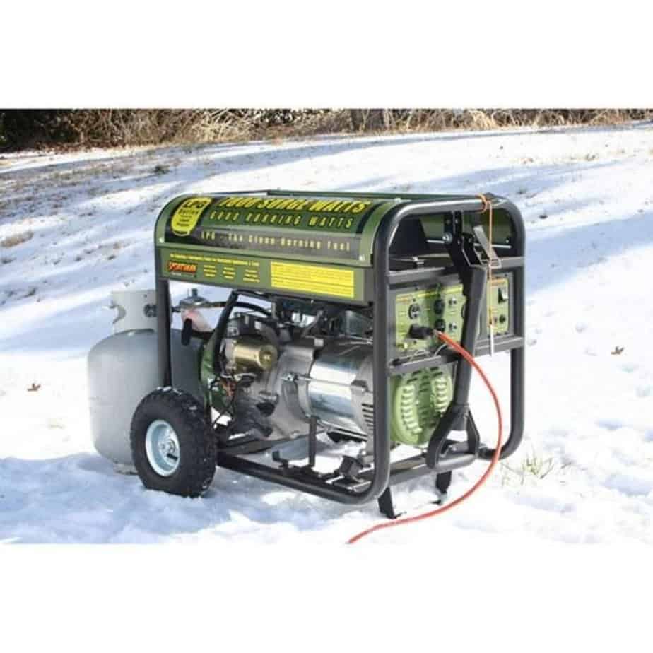 A portable generator connected to a propane tank sits in the snow with an orange extension cord running from it