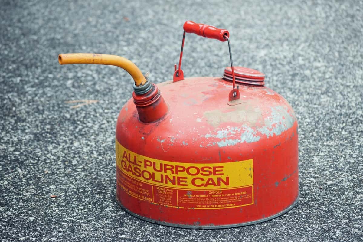 A weathered red all-purpose gasoline can with a yellow spout sitting on pavement